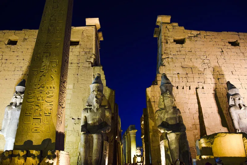 Obelisk and seated colossi at the floodlit entrance of Luxor Temple against a deep blue night sky