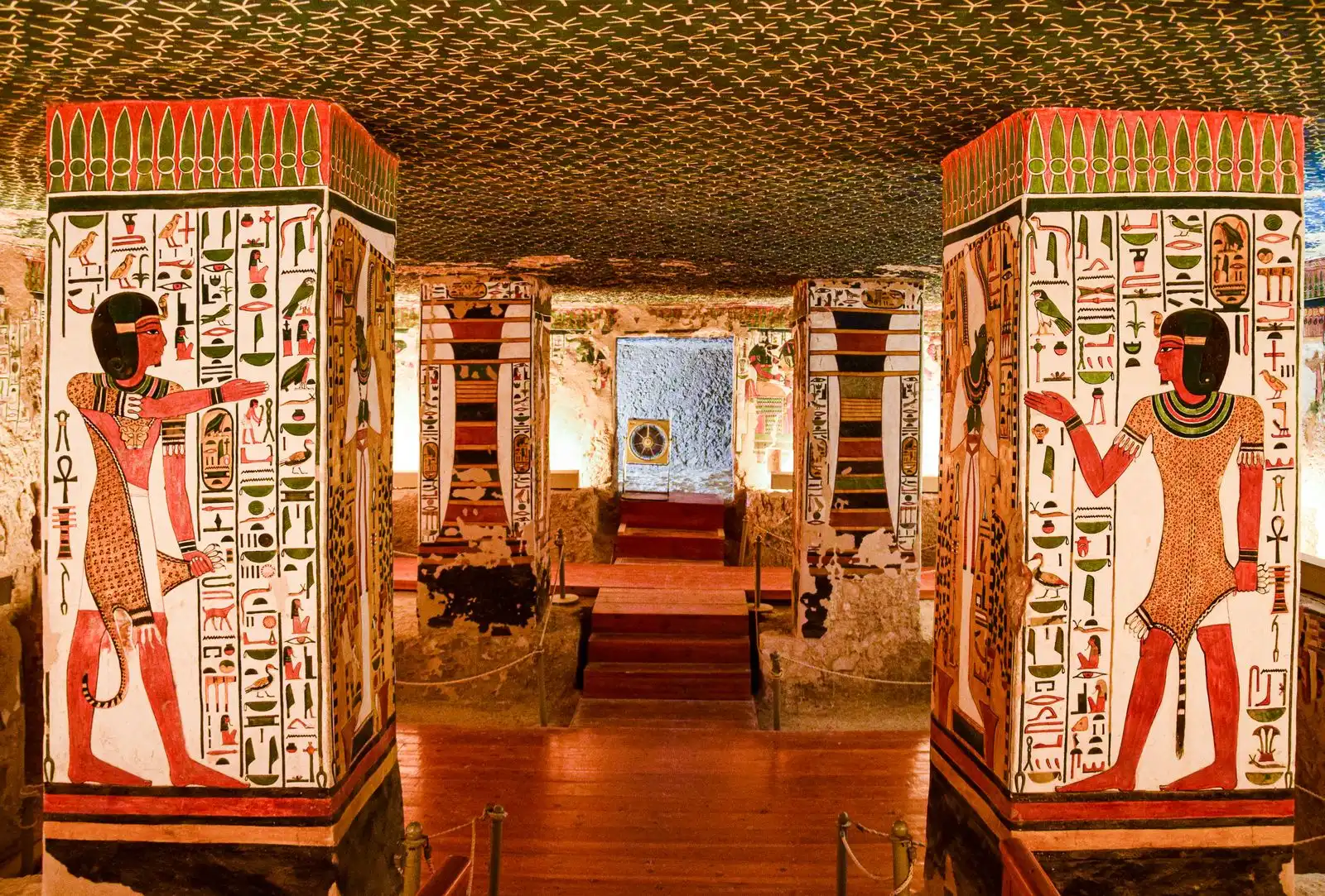 Painted pillars and walls inside a lit rock-cut tomb, with steps down to the burial chamber