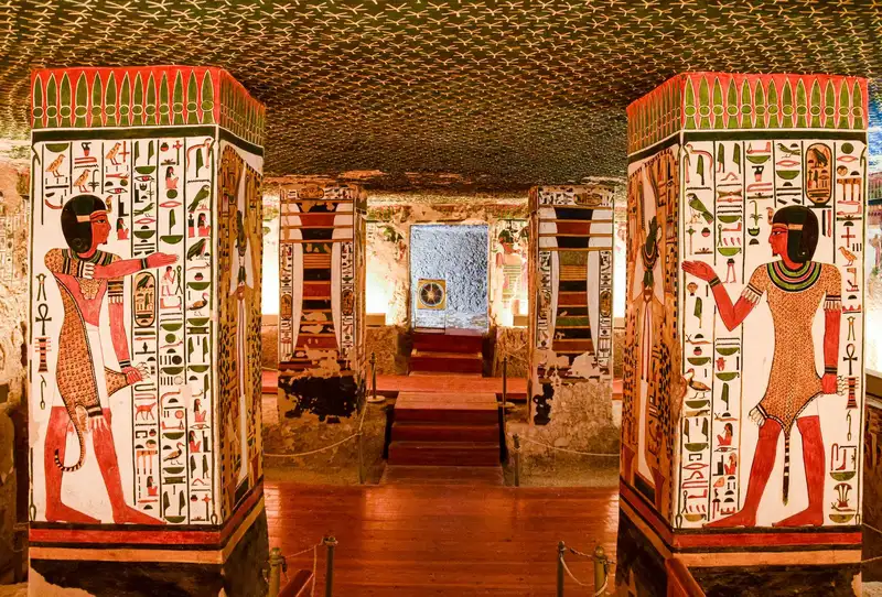 Painted pillars and walls inside a lit rock-cut tomb, with steps down to the burial chamber
