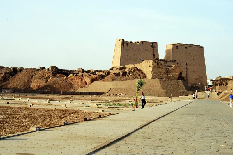 The twin pylon towers of Edfu temple seen across the paved forecourt, a young palm beside the path