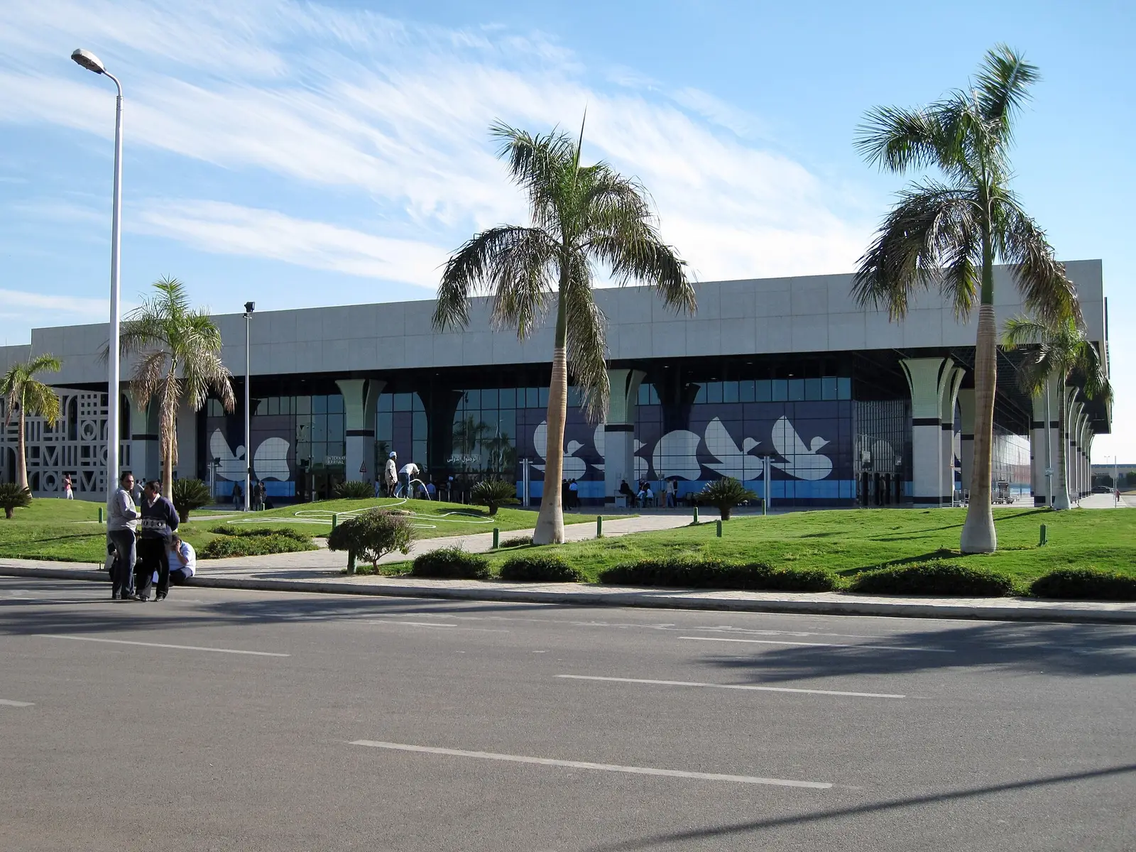 Luxor airport terminal building behind palms and a lawn, the access road in front