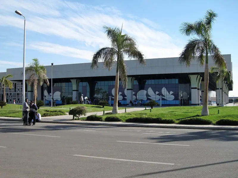 Luxor airport terminal building behind palms and a lawn, the access road in front