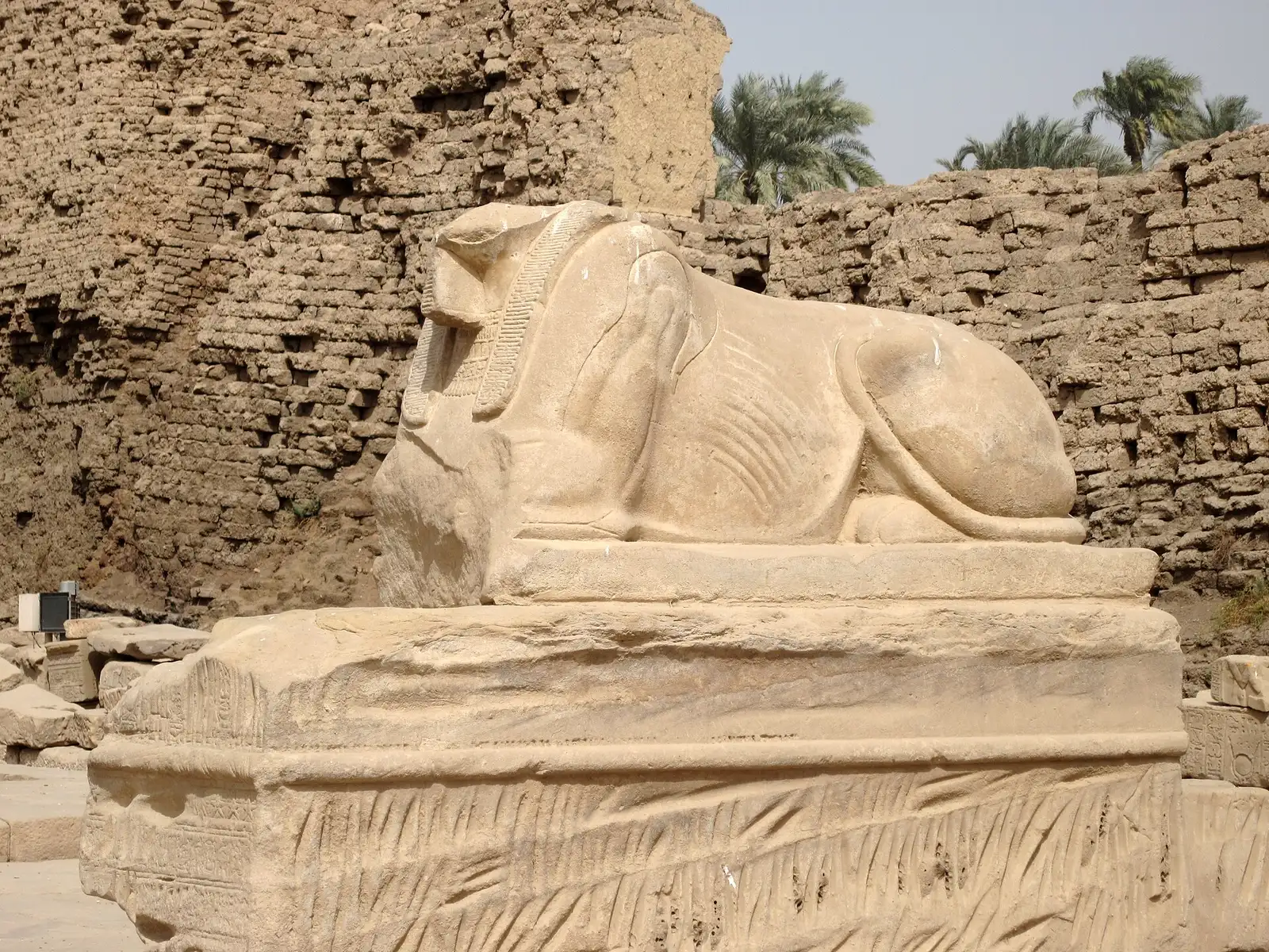 Stone sphinx on a carved plinth beside a mud-brick wall at Dendera