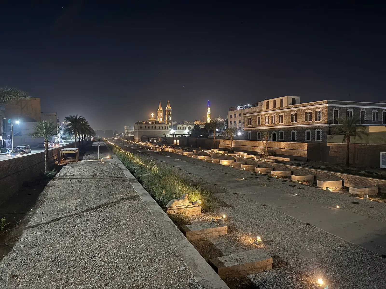 The Avenue of Sphinxes lit at night with a church and minaret beyond
