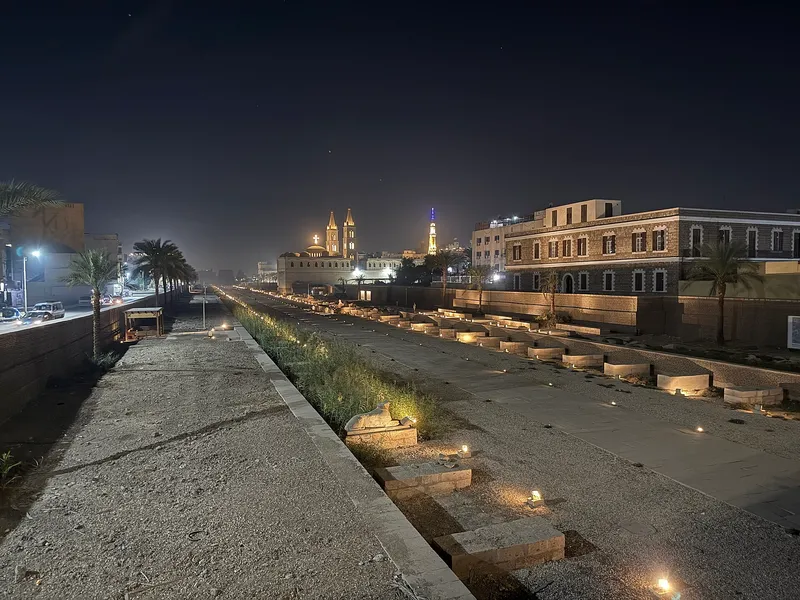 The Avenue of Sphinxes lit at night with a church and minaret beyond