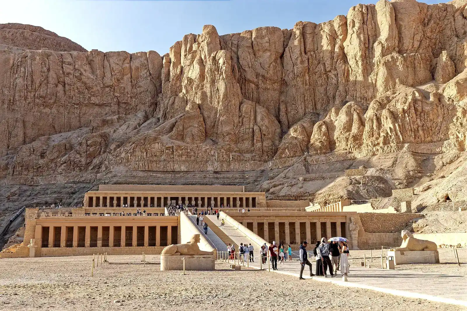 Hatshepsut's terraced temple below tall sandstone cliffs, with a sphinx on each side of the ramp and small groups of visitors in front
