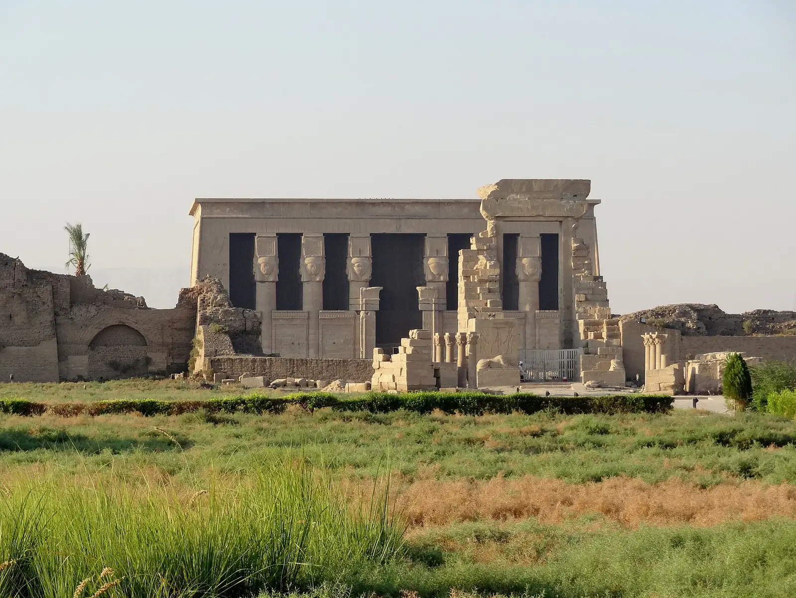 Facade of the Hathor temple at Dendera with its Hathor-headed columns, seen across green fields