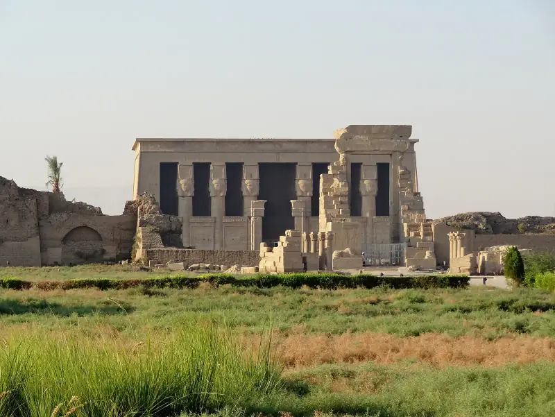 Facade of the Hathor temple at Dendera with its Hathor-headed columns, seen across green fields
