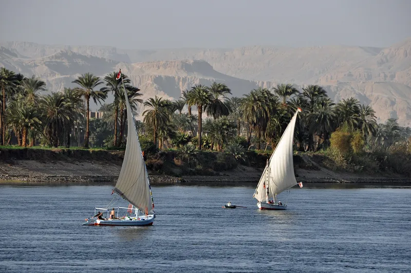 Two feluccas sailing on the Nile at Luxor, with palms and the West Bank hills behind