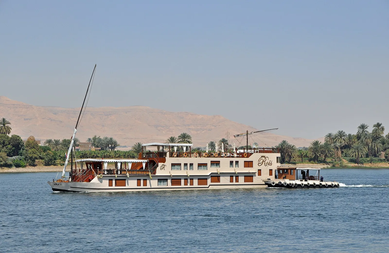 Two-masted sailing dahabiya moving along the Nile past palms, a pale desert ridge behind