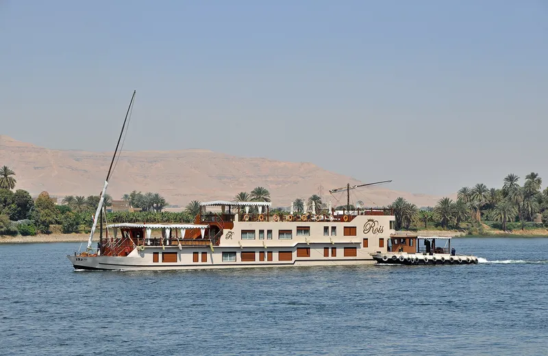 Two-masted sailing dahabiya moving along the Nile past palms, a pale desert ridge behind