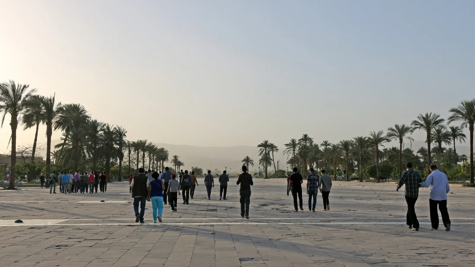 Visitors walking across the wide palm-lined entrance plaza toward Karnak in hazy morning light