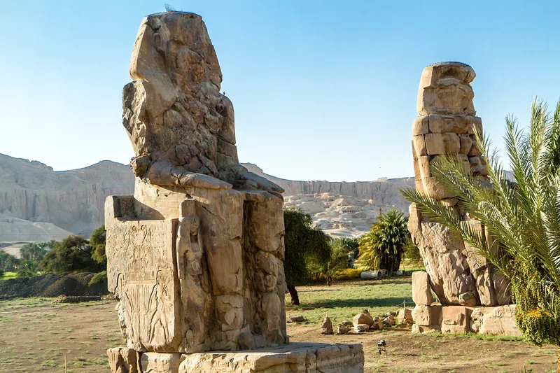 Close view of the weathered northern Colossus of Memnon, with the second statue behind palms