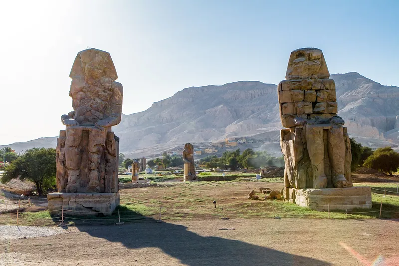 The Colossi of Memnon backlit by low sun, with the Theban hills behind