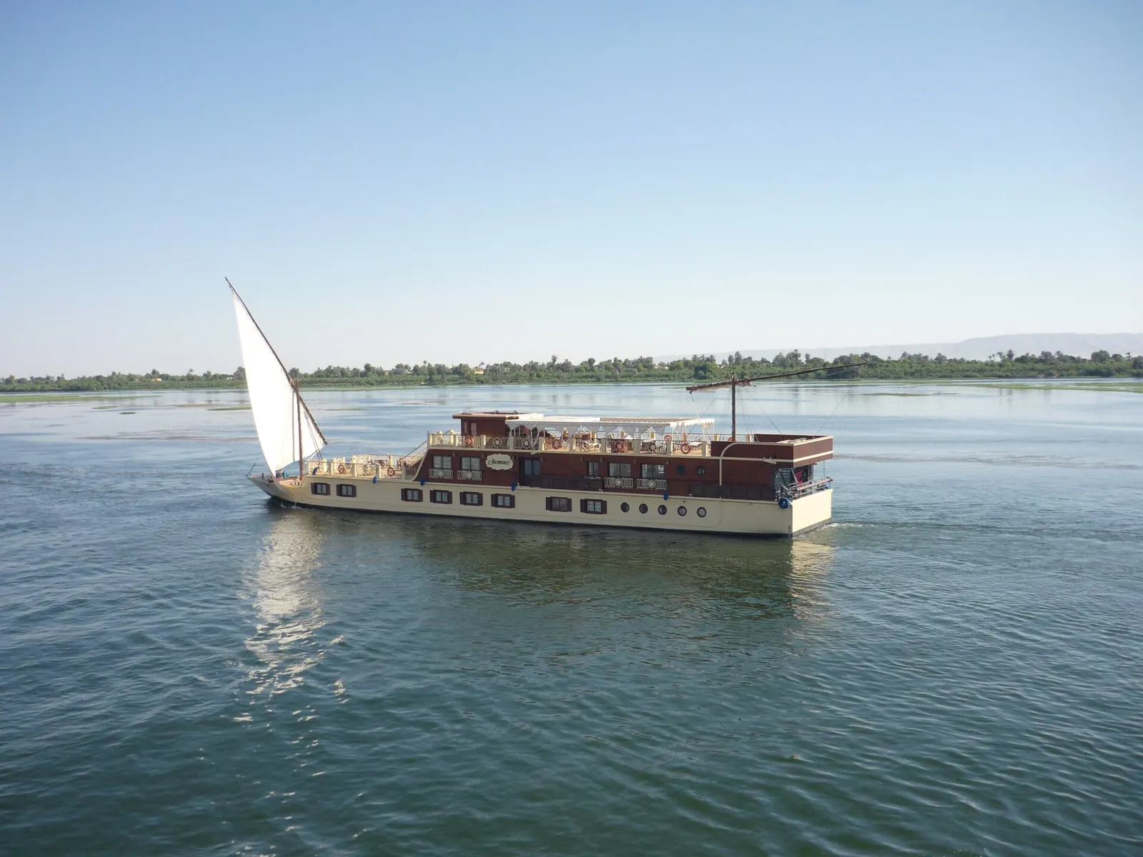 Two-deck sailing boat under a white sail on a wide, calm stretch of the Nile
