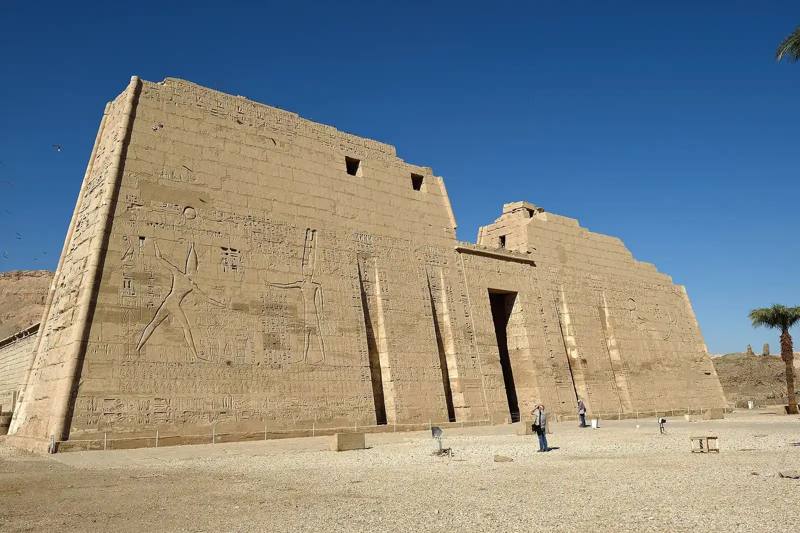 Huge carved first pylon of Medinet Habu under a deep blue sky, visitors at its foot