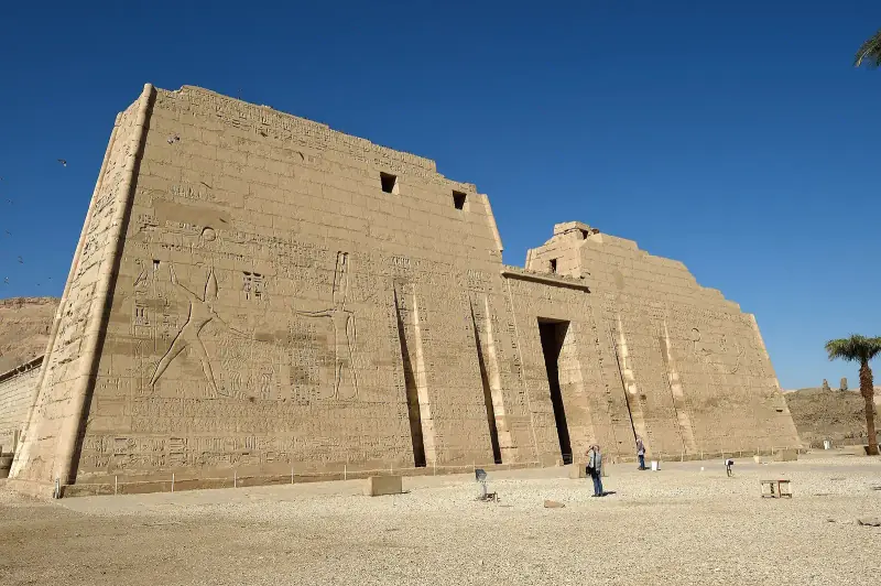 Huge carved first pylon of Medinet Habu under a deep blue sky, visitors at its foot